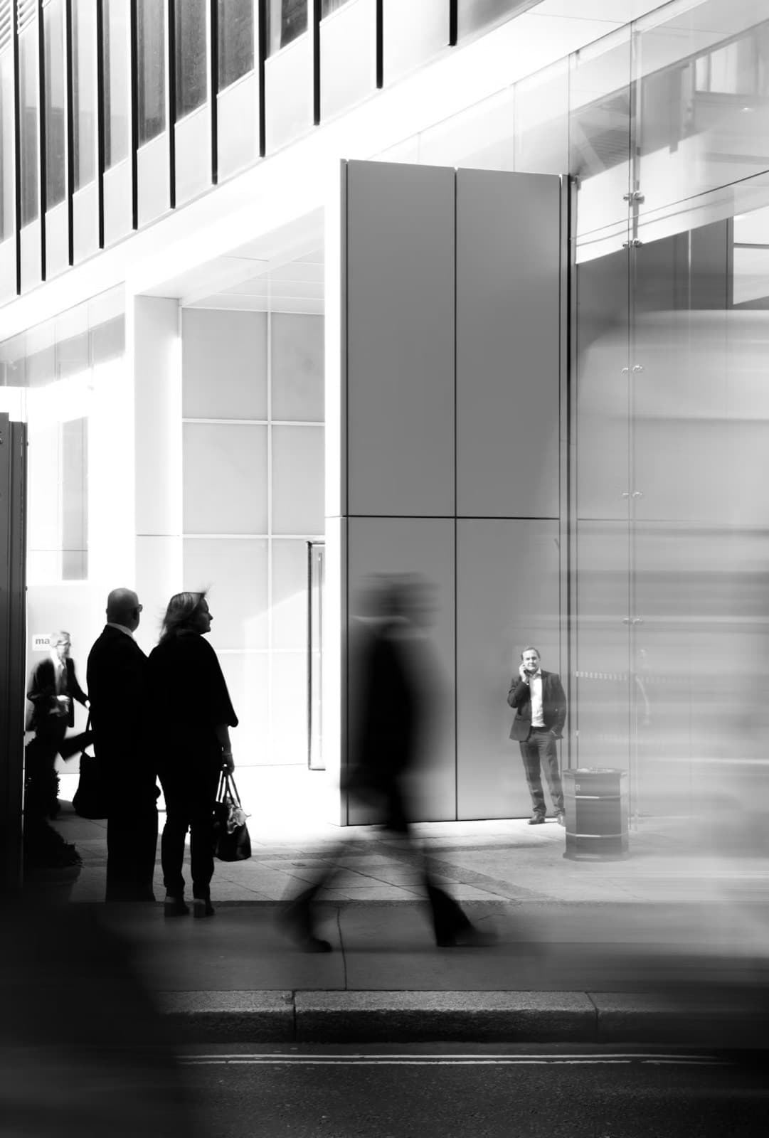 Black-and-white photograph of people crossing a bright, high-ceilinged office lobby, their figures blurred by movement